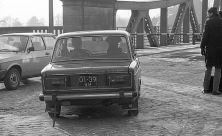Two 1970s-vintage cars and a man in woollen hat and gloves with a briefcase beside the Glienicke Bridge